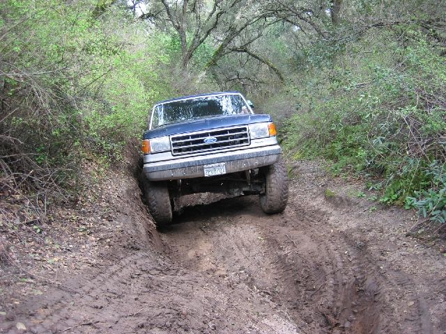 Dark blue lifted Ford Bronco descending a rutted muddy trail in the woods, front view