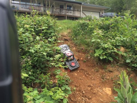 1989 Ford Bronco - A toy remote control car lies on its side on a grassy and dirt slope near a house.