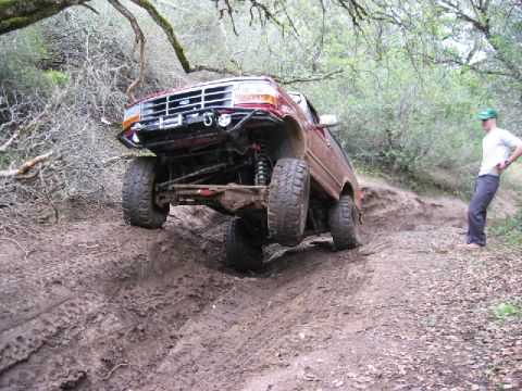 1989 Ford Bronco - A man watches as a modified Ford Bronco navigates a steep, muddy incline on a wooded trail.
