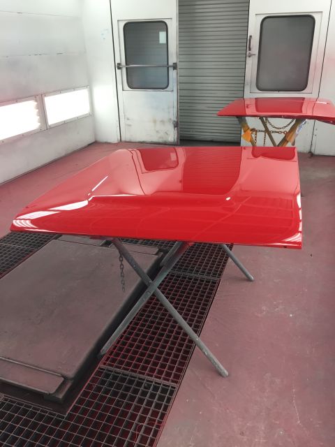 1964 Ford Fairlane - A freshly painted red car hood rests on a stand inside a paint booth.