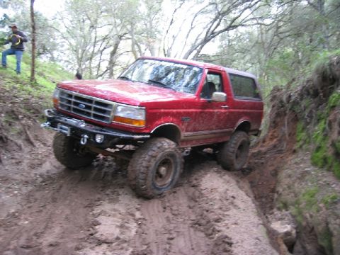 1989 Ford Bronco - A red Ford Bronco tackles a challenging muddy incline on an off-road trail.