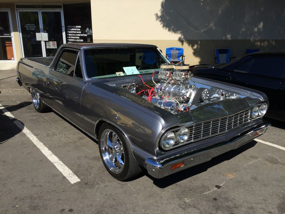 Gray 1962 Chevrolet El Camino with hood open showing a supercharged V8 engine. Parked next to a black car
