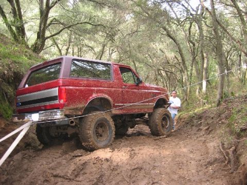 1989 Ford Bronco - A red 1989 Ford Bronco navigates a muddy off-road trail, with a man observing from the side.