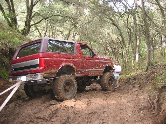 Red lifted full-size Ford Bronco on a muddy forest trail, rear 3/4, being winched