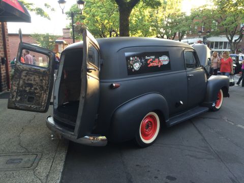 1962 Chevrolet Suburban - A matte black panel van with its rear doors ajar is parked curbside, showing off its red-rimmed wheels and a custom shop decal.