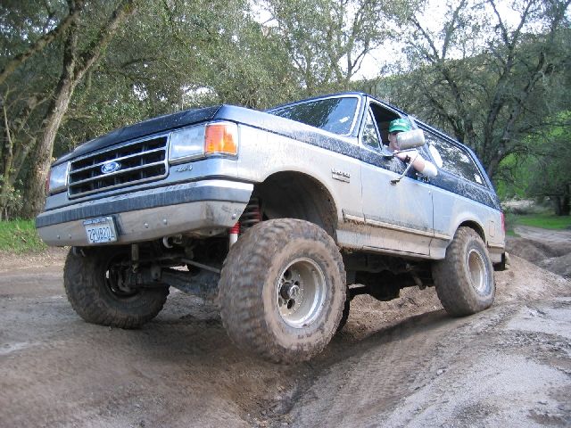 Lifted silver Ford Bronco with mud tires flexing over dirt mounds on a wooded off-road trail