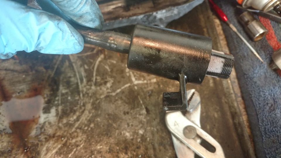 Gloved hand holds metal part with black cylindrical housing; pliers grip a bracket on the housing