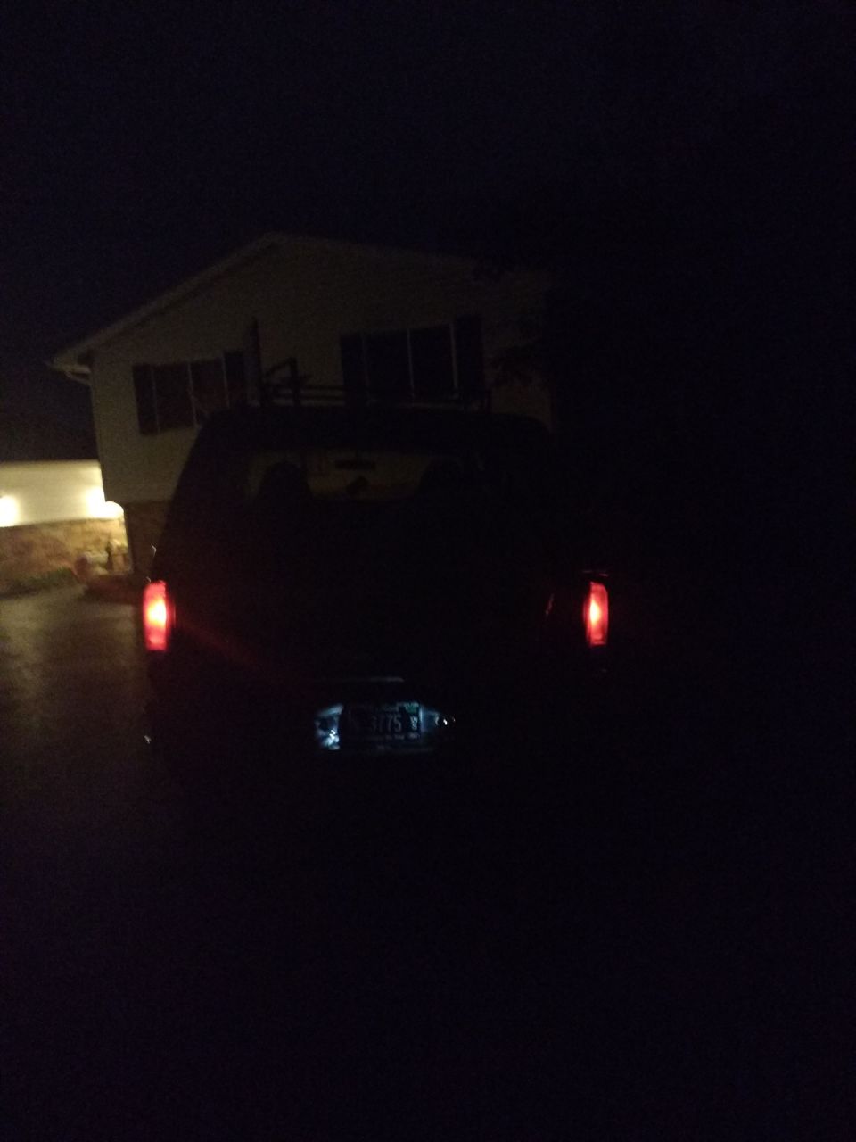 Rear view of a dark SUV at night with red taillights and lit license plate, house in background
