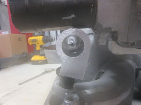 Metal bracket with circular opening, attached to a larger gray component. A yellow drill is visible in the bac