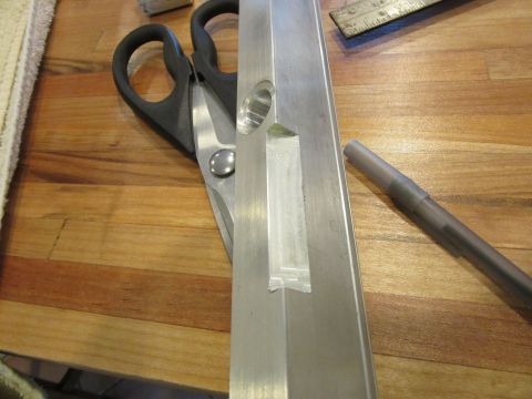 Scissors next to a metal extrusion with tape and a hole, on a wooden surface