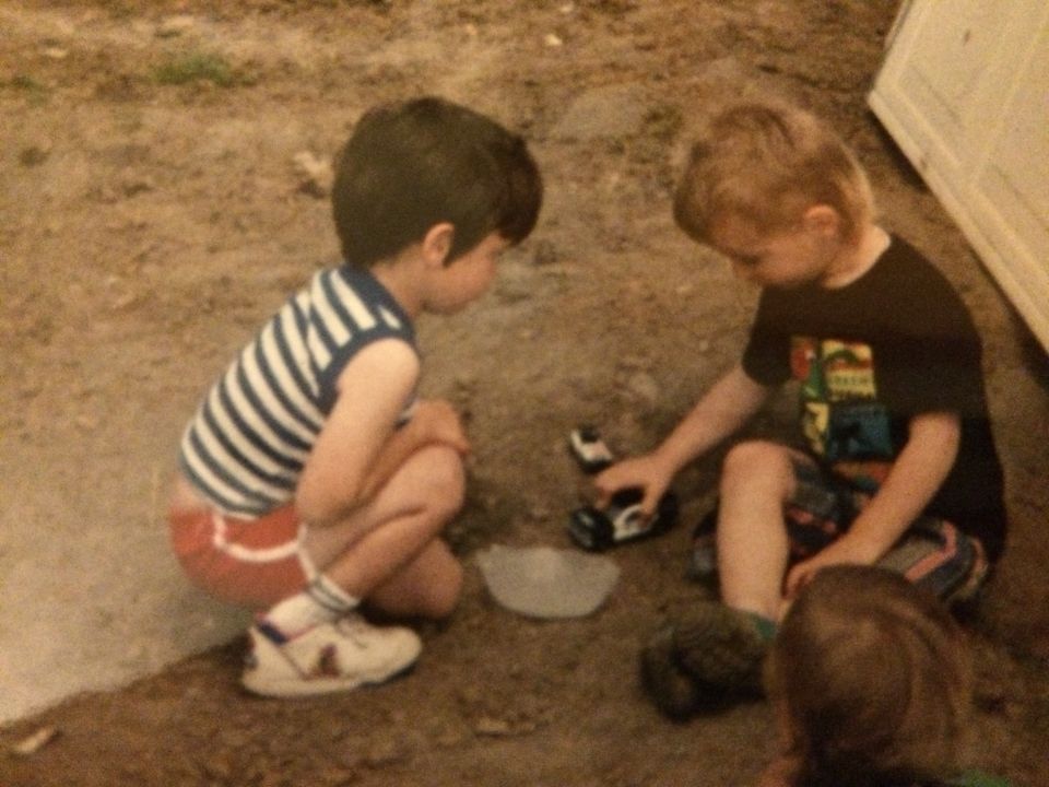 Two young boys squatting on dirt, one in a striped shirt, the other in a black t-shirt, playing with toy cars