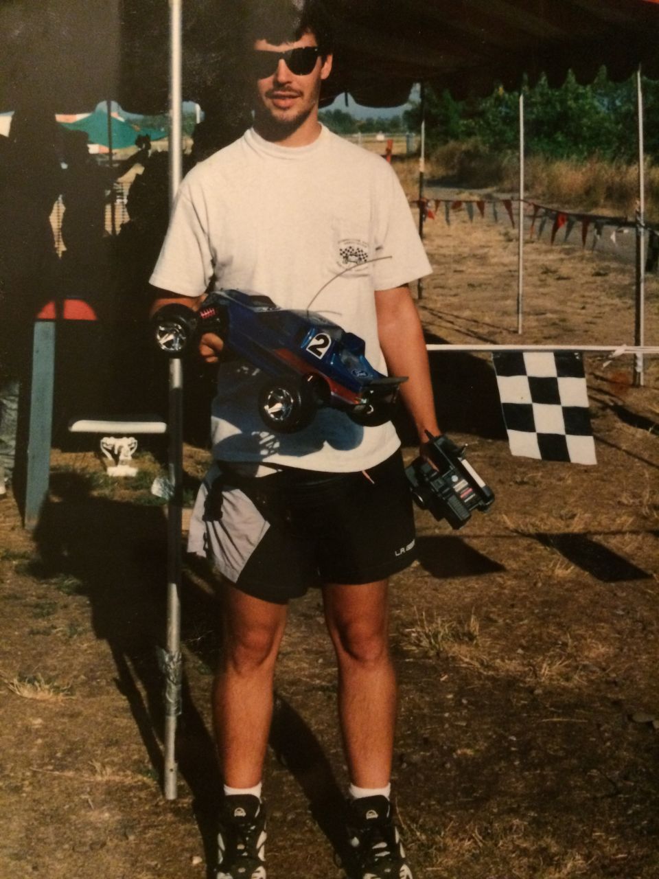 Man in t-shirt and shorts holding a blue remote control car with number 2 on it, and a remote control in his other hand. Checkered flag in background
