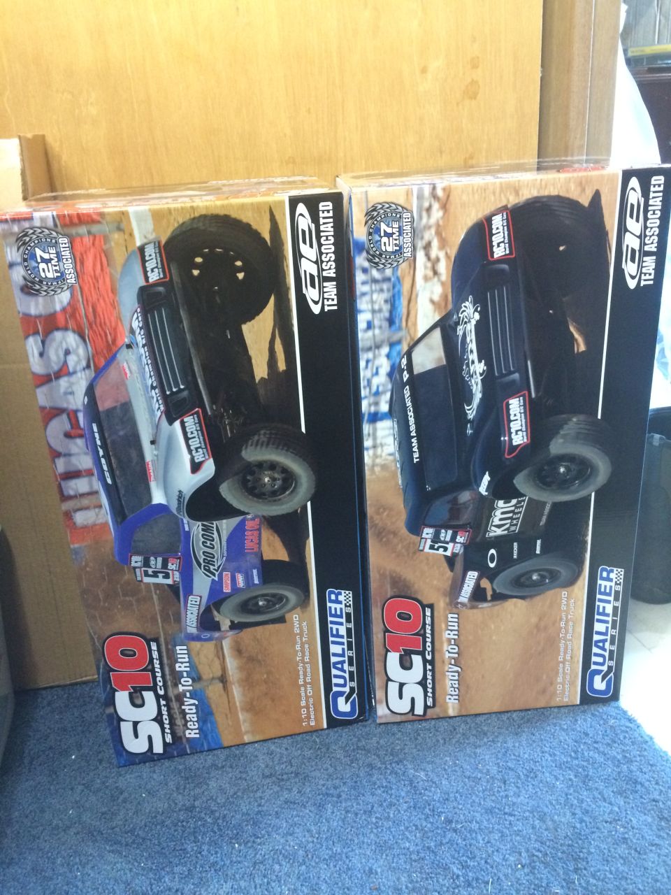 Two boxes of Team Associated SC10 Qualifier Ready-to-Run short course trucks, one blue and silver, one black, with graphics of the trucks on the boxes