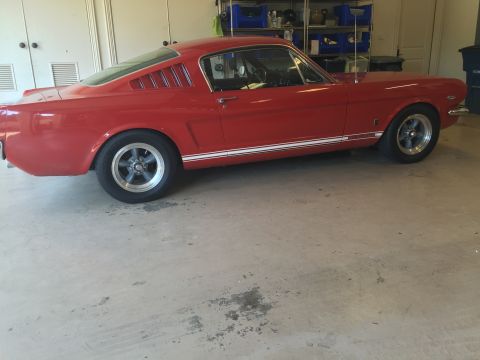 1965 Ford Mustang - A red 1965 Ford Mustang coupe is parked in a garage. The car features classic louvers on the rear quarter windows and a white stripe along…