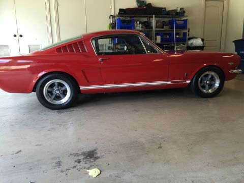 1965 Ford Mustang - A bright red 1965 Ford Mustang coupe is parked in a garage with its side facing the camera. The car features chrome trim, white racing stri…