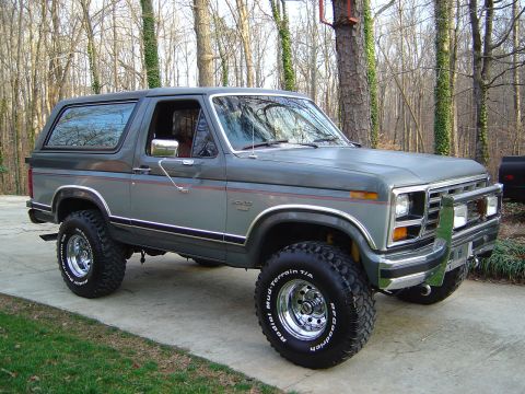 1986 Ford Bronco - A two-tone gray and dark gray 1980s Ford Bronco full-size SUV shown from a 3/4 front angle, lifted on chrome wheels with BFGoodrich Mud-Ter…