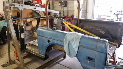 1979 Ford Bronco - A blue vehicle fender section is supported by an engine hoist and a metal stand in a garage setting. Another section of the vehicle's body,…