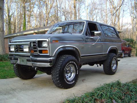 1986 Ford Bronco - A two-tone gray and silver 1980s Ford Bronco full-size SUV shown from a front three-quarter angle, lifted on BFGoodrich Mud-Terrain tires w…