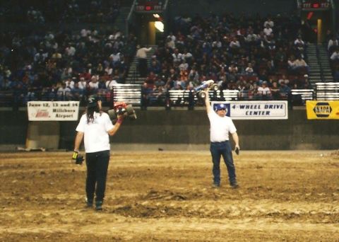 1989 Ford Bronco - Two people are holding remote-controlled monster trucks in an arena with a large crowd in the background. One person is holding a red and b…