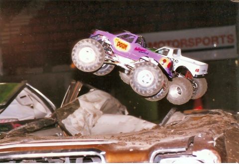 1989 Ford Bronco - A purple monster truck with the word "DOOM" on the side is airborne over a pile of crushed cars, with a white pickup truck visible behind i…