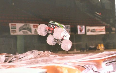 1989 Ford Bronco - A monster truck, appearing to be a Ford Bronco, is captured mid-air, jumping over debris in an arena. The truck has large, knobby tires and…