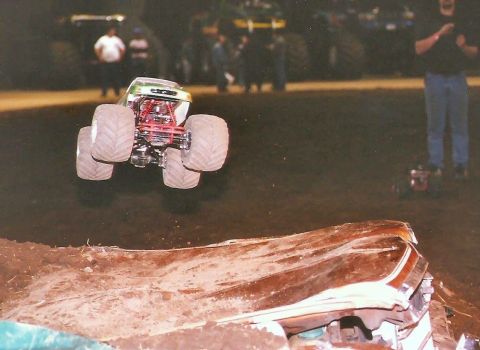 1989 Ford Bronco - A monster truck is captured mid-air, jumping over a crushed car. The truck is green and chrome with exposed red suspension components.