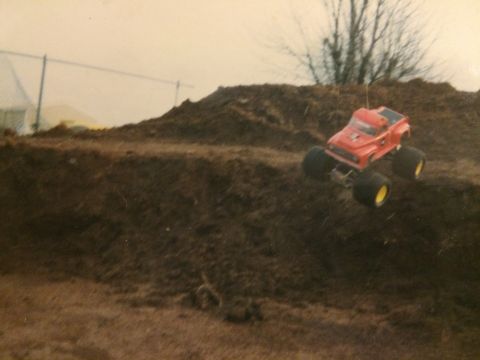1989 Ford Bronco - A red radio-controlled monster truck with yellow-rimmed wheels is captured mid-air, jumping over a dirt mound. The truck appears to be a re…