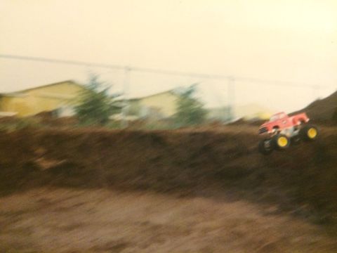 1989 Ford Bronco - A red and yellow remote-controlled monster truck is captured mid-air as it jumps over a dirt ramp. The background shows blurred buildings a…