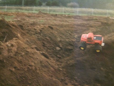 1989 Ford Bronco - A bright orange remote-controlled monster truck with large black tires and yellow rims is shown driving on a dirt hill. The truck is angled…