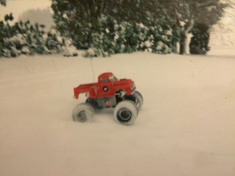 1989 Ford Bronco - A red remote-controlled monster truck with large tires is driving through fresh snow. The truck is positioned in the center-right of the fr…