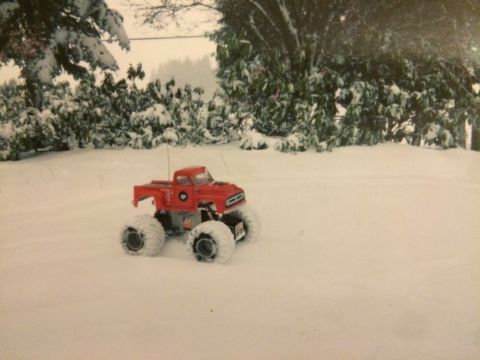 1989 Ford Bronco - A red remote-controlled monster truck with large tires sits in the snow. The truck is positioned in a snowy outdoor setting with trees and…