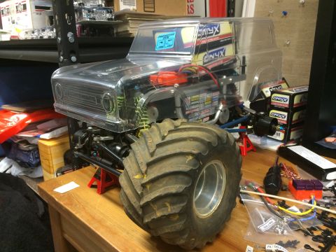 1989 Ford Bronco - A clear plastic body of a monster truck is shown on a wooden table, with a large tire in the foreground and various RC car parts scattered…