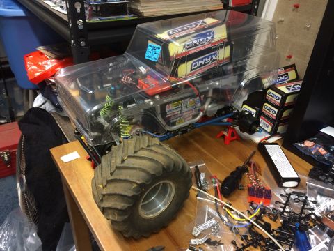 1989 Ford Bronco - A clear-bodied RC monster truck with large tires and "ONYX" branded batteries is assembled on a wooden table amidst various parts and tools.