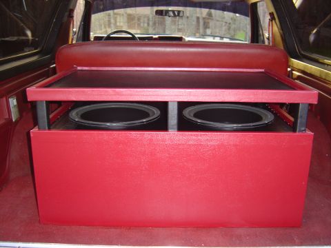 1986 Ford Bronco - Interior rear cargo area of a vehicle showing a custom red vinyl-wrapped subwoofer box housing two downward-facing speakers under a lift-up…
