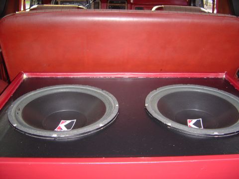 1986 Ford Bronco - A carpeted subwoofer enclosure holding two Kicker 12-inch speakers, mounted behind a red vinyl rear seat inside a vehicle interior.