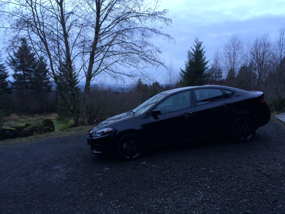 Black 2015 Dodge Dart sedan parked on a gravel driveway with trees in the background.