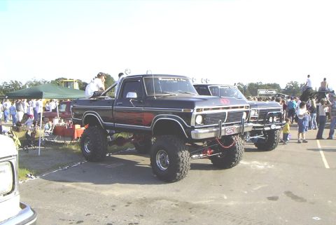 1995 Ford Bronco - A lifted pickup truck with flame decals is parked at an outdoor event with other vehicles and people.