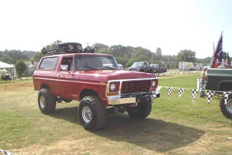 1995 Ford Bronco - This lifted red Ford Bronco features a roof rack with a spare tire and auxiliary lights, parked at an outdoor event.