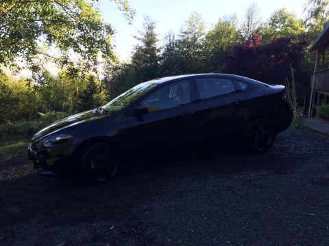 2015 Dodge Dart - A black 2015 Dodge Dart sedan is parked on a gravel driveway next to a wooded area with a house visible on the right.
