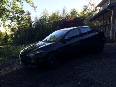 2015 Dodge Dart - A black sedan is parked on a gravel driveway in front of a house with a porch. The car is angled towards the front left, with trees and fol…