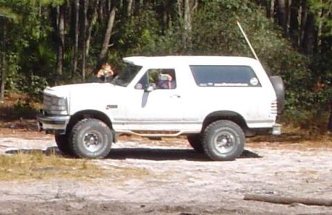 1996 Ford Bronco - Side view of a white 1996 Ford Bronco parked on a dirt road surrounded by trees.