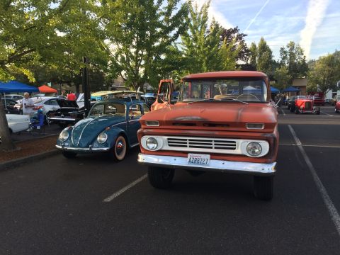 1962 Chevrolet Suburban - A teal blue Volkswagen Beetle and an orange Chevrolet pickup truck are parked side-by-side in a parking lot with trees in the background.