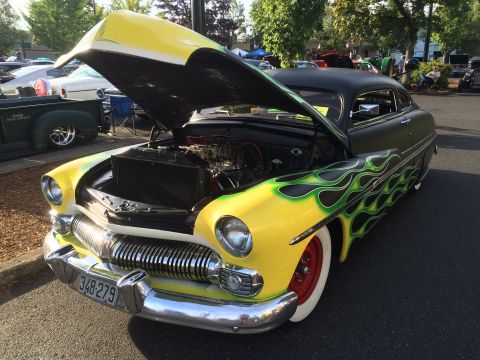 1962 Chevrolet Suburban - A custom yellow and black 1950s era coupe with green flame graphics is parked outside with its hood open, revealing the engine. The car fea…