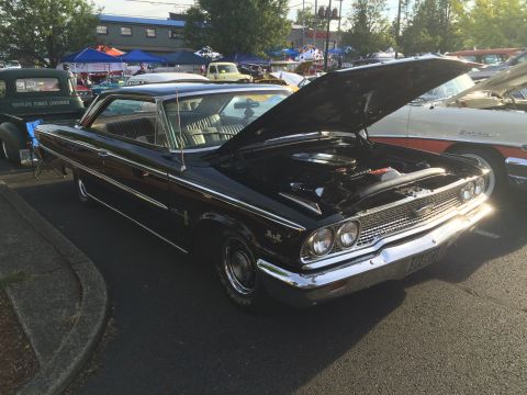 1962 Chevrolet Suburban - A black 1960s era coupe with its hood open is parked in an asphalt lot next to a green pickup truck. The classic car has chrome accents and…
