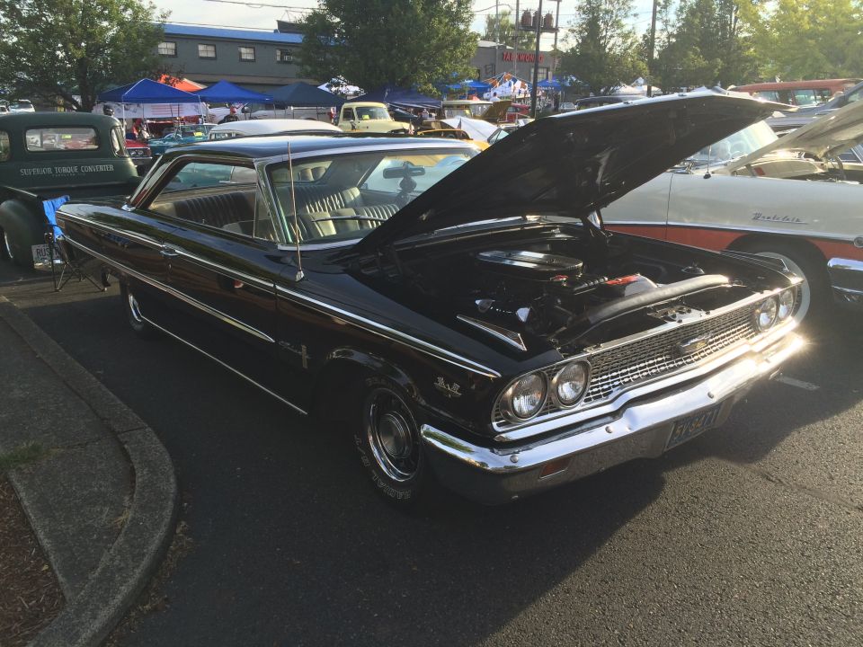 Black 1960s coupe with hood open, chrome grille, and visible engine, parked next to a green pickup truck.
