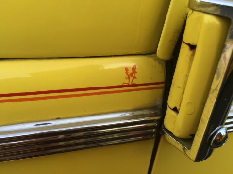 1962 Chevrolet Suburban - A close-up view of the side of a yellow vehicle shows a small red graphic of two figures and three horizontal stripes. A chrome trim piece…