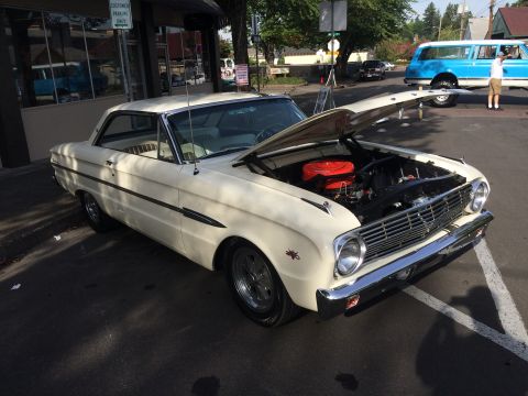 1962 Chevrolet Suburban - A cream-colored 1960s coupe with its hood open, revealing the engine, is parked on an asphalt surface in front of a building. A blue SUV is…