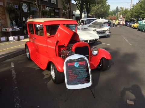 1962 Chevrolet Suburban - A red 1928 Ford Model A sedan is parked on a street next to other vehicles and shops. A sign in front of the car lists details about its bo…