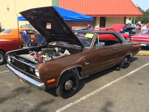 1962 Chevrolet Suburban - A brown 1960s coupe with its hood open, revealing a V8 engine, is parked outdoors at a car show. Other vehicles and a building with a red r…
