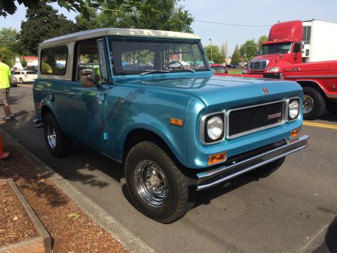 1962 Chevrolet Suburban - A bright teal 1960s International Harvester Scout II is parked on the side of the road, with a red semi-truck visible in the background. Th…